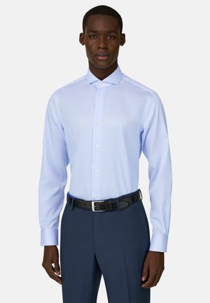 Mann trägt hellblau gestreiftes Hemd, in marineblaue Anzughose mit schwarzem Gürtel gesteckt, steht vor schlichtem weißem Hintergrund.