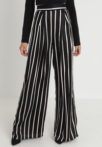 Femme portant un pantalon noir taille haute à jambes larges avec des rayures blanches verticales et un haut noir à manches longues, debout devant un fond uni.