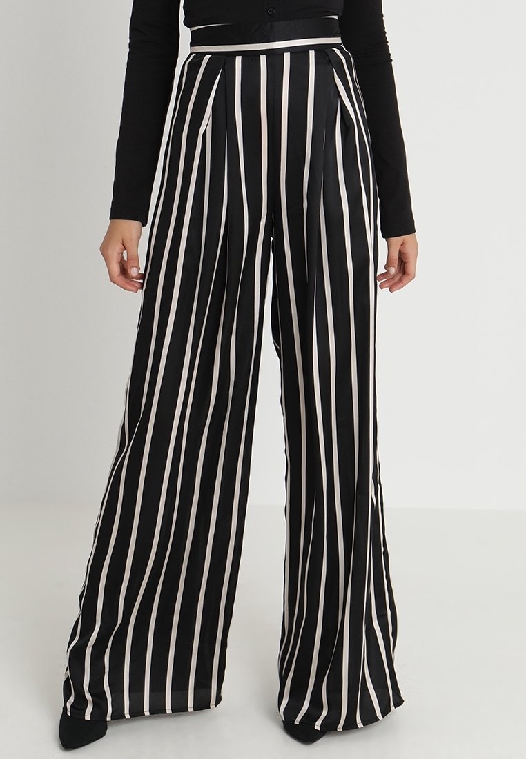 Femme portant un pantalon noir taille haute à jambes larges avec des rayures blanches verticales et un haut noir à manches longues, debout devant un fond uni.