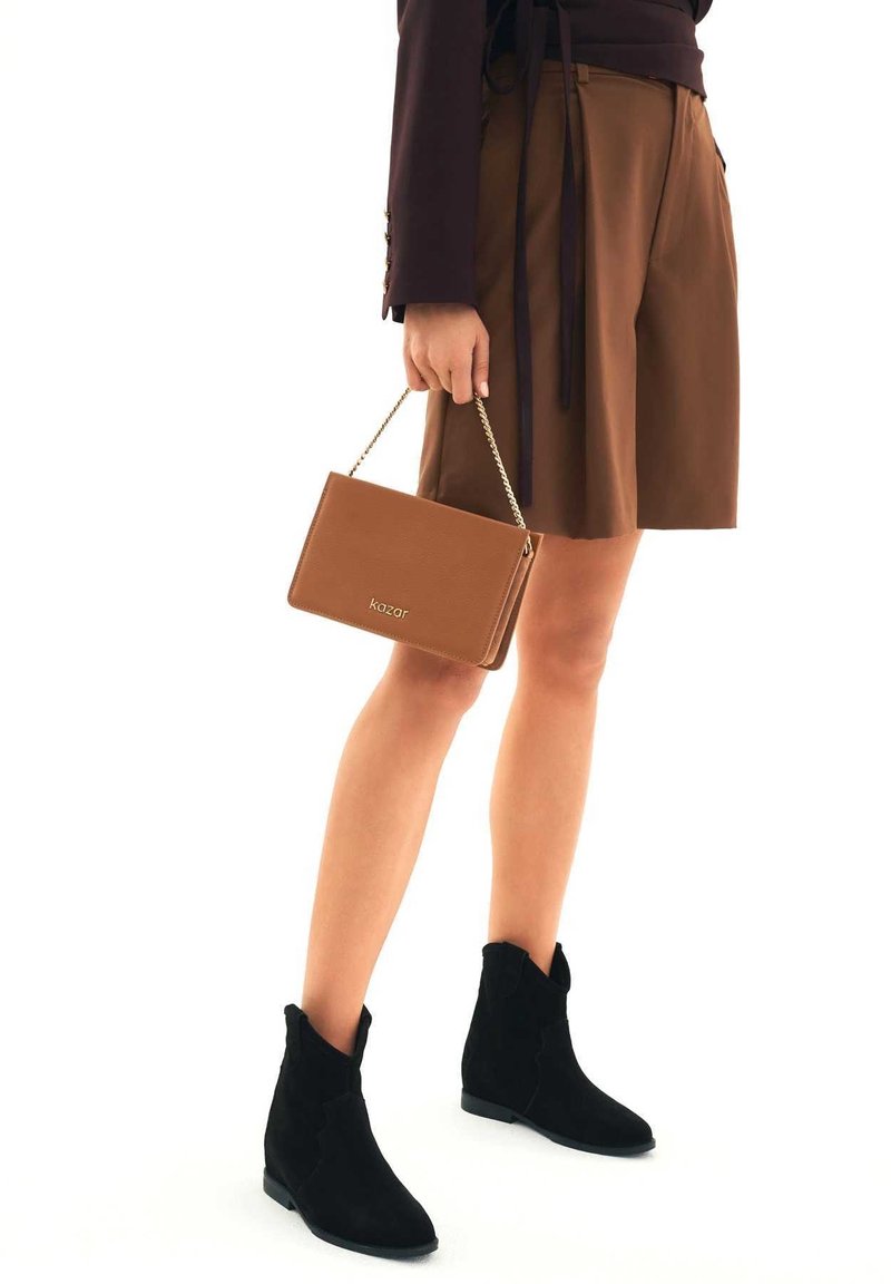 Nogi kobiety w czarnych zamszowych botkach do kostki, brązowej plisowanej spódnicy, trzymającej beżową skórzaną torebkę z złotym łańcuszkiem.