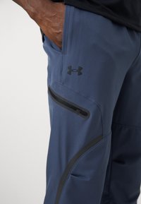 Pantalons de sport navy en matériau flexible avec une poche latérale zippée et des accents noirs ; un logo Under Armour est visible sur la cuisse.