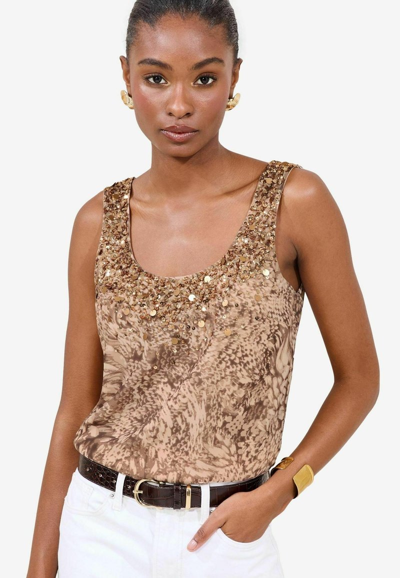 Sleeveless top in beige with a textured pattern, embellished with gold sequins around the neckline. Paired with a dark belt and white pants.
