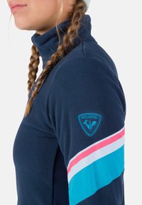 Námořnický fleecový pulovr s vysokým límcem, který má na rukávu jasně modrý a růžový pruhovaný design a logo nášivku na rameni.