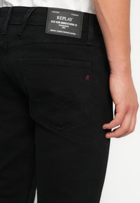Černé džíny z denimu mají hladkou texturu, decentní prošívání a logovou nášivku na pase. Zadní kapsa obsahuje červený vyšívaný detail.