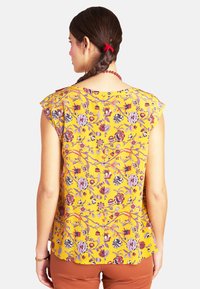 Gelbe Bluse mit Blumenmuster und Kappärmeln, verziert mit lila und roten Mustern. Leichter Stoff, runder Ausschnitt und gerader Schnitt.