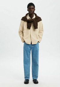 Ung mand iført en beige skjorte med knapper, blå jeans, sorte sko og en mørkebrun sweater bundet om skuldrene.