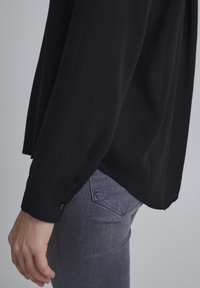 Zwarte langarmshirt met een gladde textuur en knoopmanchetten, gecombineerd met slim-fit grijze jeans met achterzakken.