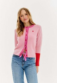 Różowy kardigan z długimi rękawami, czerwonymi akcentami, z zapięciem na guziki i małym detalem w kształcie serca. Stylizowany z dżinsami o wysokim stanie i związana różową wstążką.