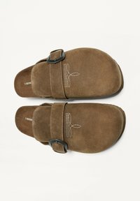 Rjave suede klompe z nastavljivim črnim sponko, zaobljenim prstom in belim šivom na zgornji površini. Ravna podplata.