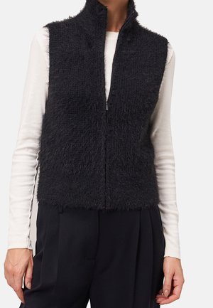 Frau trägt eine schwarze, flauschige ärmellose Zip-Weste über einem weißen Langarmshirt und schwarzen Hosen vor schlichtem Hintergrund.