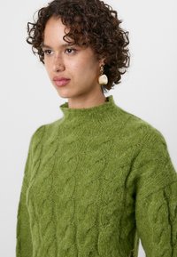Pull en tricot vert avec un col haut, arborant un motif en relief de câbles. Un modèle aux cheveux bouclés porte de larges boucles d'oreilles en ton doré.