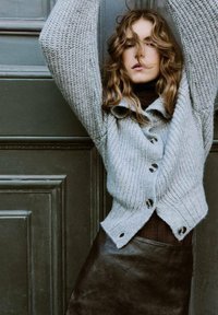 Cárdigan de punto gris claro con textura acanalada y grandes botones, combinado con una falda de cuero marrón oscuro. El fondo es de color verde oscuro.