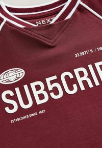 Burgundia puuvillane jersey valge äärega. Omab "SUB5CRIP" teksti ja maakera logo. Kaasas on koordinaadid ja tekst "KÄIVITATUD ALATES. 1982."