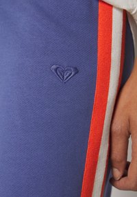 Paarse sportieve broek met rode en witte zijstrepen. Voorzien van een geborduurd hartlogo op de dij. Zachte, gebreide textuur.