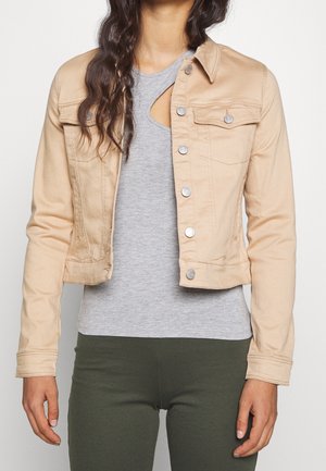 Vrouw draagt een beige cropped jas over een grijze top met een zijuitsnijding en donkergroene nauwsluitende broek, staand tegen een witte achtergrond.