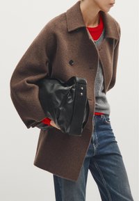 Bruine wollen jas met een kraag, twee zwarte knopen en een relaxte pasvorm. Het model houdt een zwarte leren tas met zilverkleurige accessoires vast. Rood en grijs in lagen eronder.