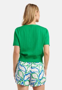 Grüner, gestrickter Kurzarmpullover mit einem strukturierten Muster, kombiniert mit weißen Shorts, die ein blau-grünes Blumenmuster zeigen.