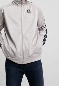 Lys grå zip-up jakke med høj krave, lavet af glat stof. Har kontrasterende sorte sidelister med logo og ribbet kant.