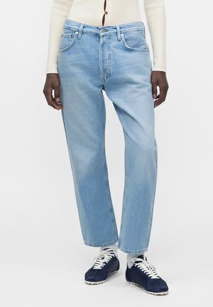Person iført lyseblå jeans med lige ben, cremefarvet ribstrikket cardigan, hvide sokker og marineblå sneakers, stående mod en hvid baggrund.