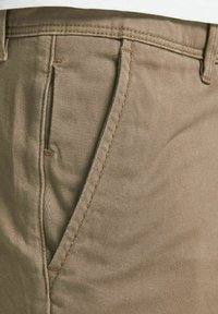 Beige kalhoty s hladkým, texturovaným materiálem; mají zúžený střih, zakřivené otvory na kapsy a kontrastní hnědé stehy.