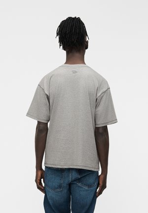 Persoon met een donkere huid en dreadlocks, die een eenvoudig grijs T-shirt met korte mouwen en blauwe spijkerbroek draagt, gezien van achteren tegen een effen achtergrond.