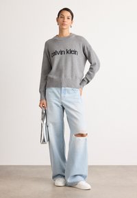Suéter gris de punto con el logo negro de "Calvin Klein", combinado con unos pantalones de mezclilla anchos claros y desgastados, y zapatillas blancas, sosteniendo un clutch plateado.