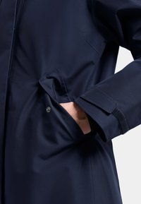 Marint vattentålig jacka med en slät yta, sidofickor och justerbara ärmslut med kardborrestängning, som framhäver funktionella designelement.