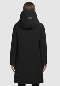 Henkilö, jolla on pitkä musta huppupäällinen talvitakki, selkä kameraan päin, ja takin hupussa sekä alaosassa on "KHUJO"-brändäys.