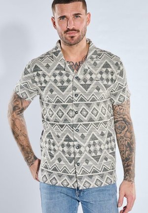 Man with tattoos wearing black-and-white geometric patterned short-sleeve button-up shirt and blue jeans, standing with one hand on hip.