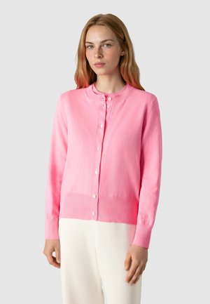 Jonge vrouw met een lichte huid en lang blond haar, gekleed in een roze vest met knopen en een witte broek, staand tegen een grijze achtergrond.