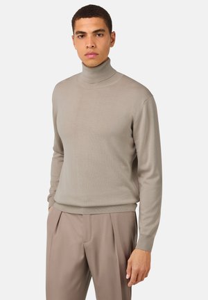 Pull en col roulé beige en tricot doux, doté d'une silhouette ajustée et de poignets et bas côtelés, associé à un pantalon clair.