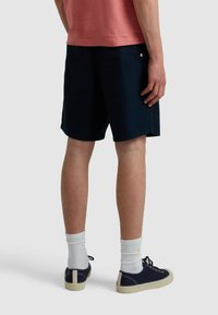Marineblaue, knielange Shorts mit glatter Textur, zwei seitlichen Taschen und dezentem Branding. Kombiniert mit weißen Socken und dunklen Schuhen.