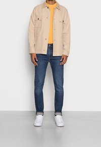 Hombre con chaqueta beige de botones, camisa amarilla, jeans azules con puños y zapatillas blancas, de pie contra un fondo blanco liso.