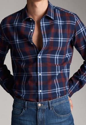Homme portant une chemise à carreaux bleu marine, marron et bleu clair, partiellement déboutonnée, rentrée dans un jean bleu, les mains dans le dos.