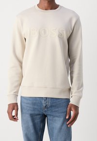 Krämfärgad sweatshirt med rund halsringning, ribbad muddar och en strukturerad "BOSS"-logotyp över bröstet. Matcher med blåjeans.