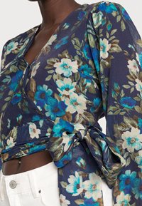 Haut cache-cœur floral de couleur bleu marine, avec des fleurs bleues et crème. Le tissu présente une finition texturée et un détail noué à la taille.