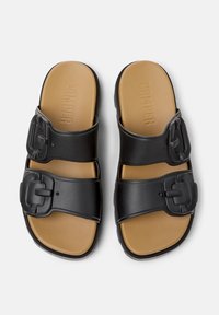 Black slide sandals with two wide straps featuring large buckles, textured rubber sole, and a smooth, tan insole with embossed logo.