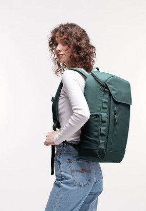 Grüner Textilrucksack mit strukturierter Form, oberem Umschlag, seitlichem Reißverschluss und verstellbaren Trägern, getragen auf dem Rücken einer Person in lässiger Kleidung.