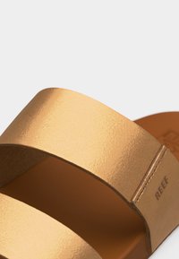 Καφέ σανδάλι με δύο λουριά, λεία υφή και ορατές ραφές, με το ανάγλυφο όνομα της μάρκας "REEF" στο πλαϊνό λουράκι.