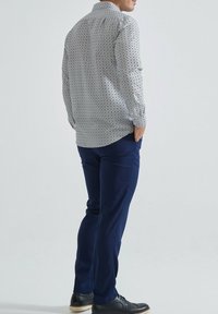 Homme debout de côté avec les mains dans les poches, portant un pantalon bleu foncé, des chaussures noires et une chemise à manches longues blanche à motifs.