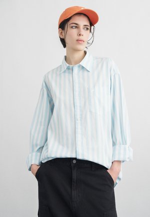 Inimene oranži mütsi, hele sinise ja valge triibulise nööbitava särgi ning mustade pükstega, käed taskus, küljele vaadates.