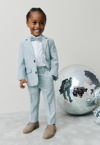 Nasmejan otrok v svetlo modrem kostimu, beli srajci in metuljčku stoji poleg velikih odsevnih disko krogel na sivi tleh ob običajni steni.