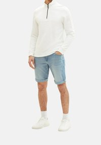 Jersey de cuello con cremallera blanco, pantalones cortos de mezclilla azul claro con dobladillos enrollados, zapatillas blancas y calcetines blancos hasta el tobillo, de pie sobre un fondo neutral.