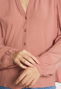 Chemisier rose à boutons avec des manches longues froncées. Tissu lisse, décolleté en V profond, et quatre boutons assortis sur le devant.