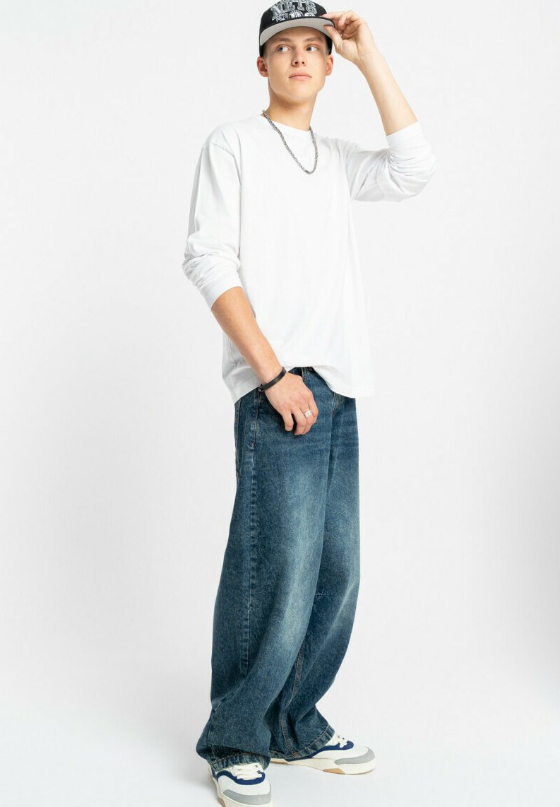 Biała koszula z długim rękawem w połączeniu z luźnymi niebieskimi dżinsami. Czapka w kolorze czarnym z białymi akcentami oraz białe sneakersy z niebieskimi detalami dopełniają stylizację.