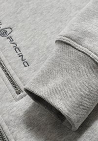 Sudadera gris con cremallera en la parte delantera y puños acanalados. La tela tiene un acabado texturizado con costuras oscuras y detalle del logotipo.