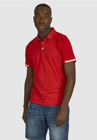 Rode polo met een kraag, een placket met twee knopen en contrasterende witte en rode gestreepte manchetten. Model draagt denim shorts.
