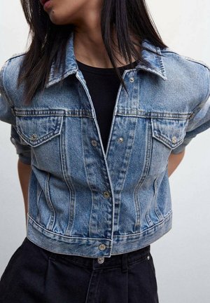 Femme portant une veste en denim bleu clair par-dessus un chemisier noir, avec de longs cheveux foncés, debout devant un fond clair uni.