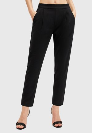 Femme portant un pantalon noir taille haute, fuselé, avec poches latérales et des talons ouverts noirs sur un fond uni.