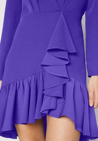 Lila klänning med långa ärmar, en volang-asymmetrisk design på sidan och en utsvängd nederkant med lager av volanger.